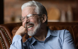 Older man with gray hair and glasses smiling and showing off dental implants