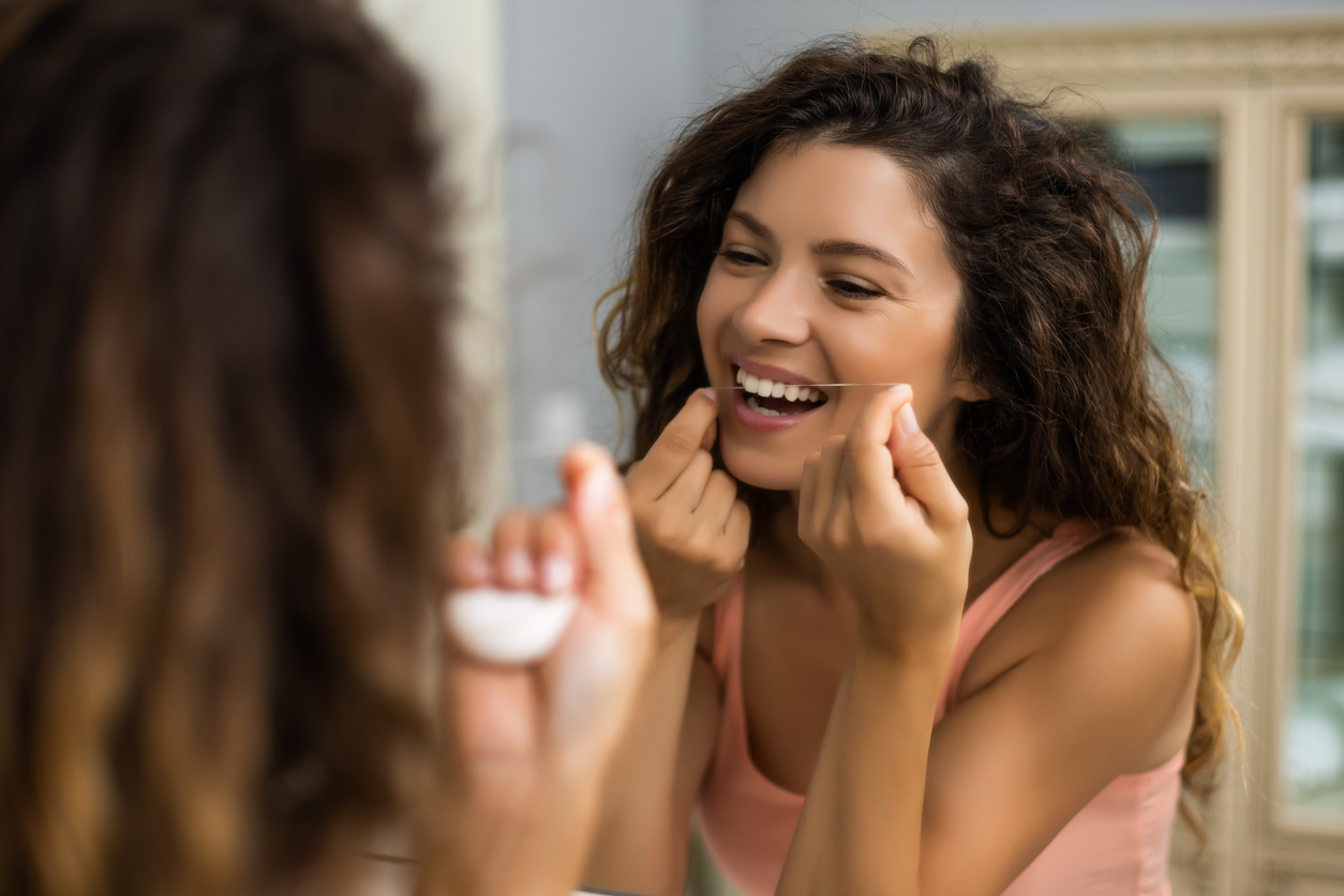 A young woman flossing her teeth in the bathroom mirror after learning about the different reasons to floss everyday - Creekview Dental