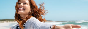 Happy mature woman with arms outstretched feeling the breeze at beach. Beautiful middle aged woman with red hair and arms up dancing on beach in summer during holiday. Mid lady in casual feeling good and enjoying freedom with open hands at sea, copy space.