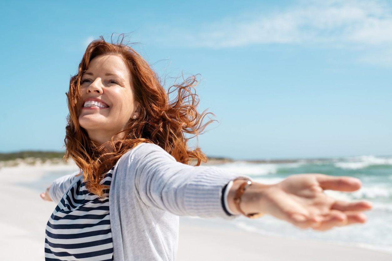 Happy mature woman with arms outstretched feeling the breeze at beach. Beautiful middle aged woman with red hair and arms up dancing on beach in summer during holiday. Mid lady in casual feeling good and enjoying freedom with open hands at sea, copy space.